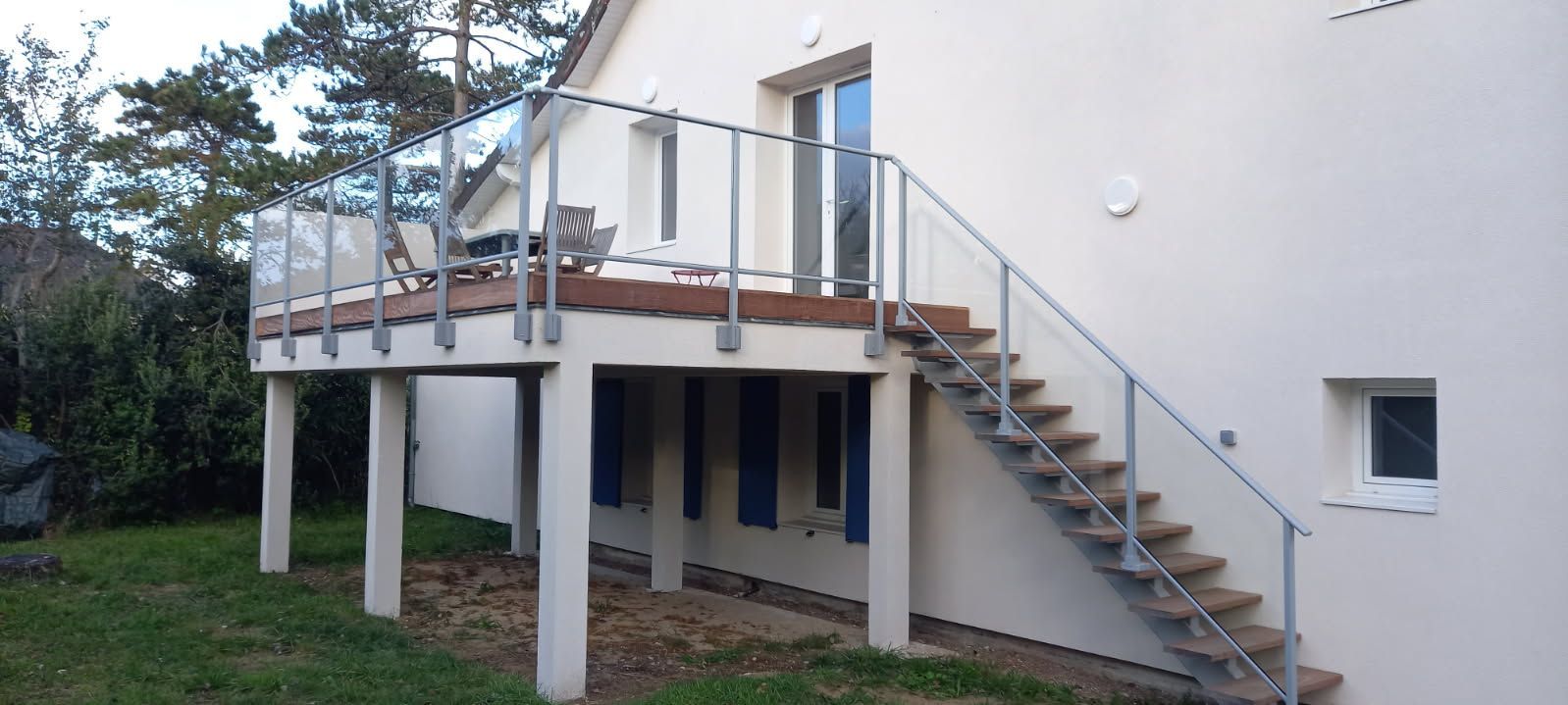 Une maison avec une terrasse en bois et un escalier extérieur. La terrasse est dotée de garde-corps en verre.