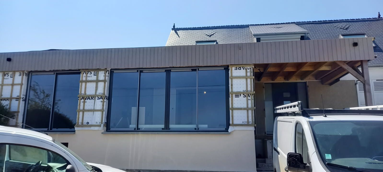 Bâtiment en construction avec de grandes fenêtres, un revêtement en bois et des véhicules stationnés.