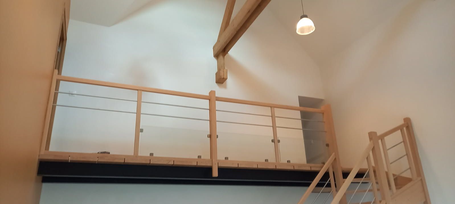Vue intérieure d'un loft avec rampe et escalier en bois. Un luminaire est suspendu au plafond.