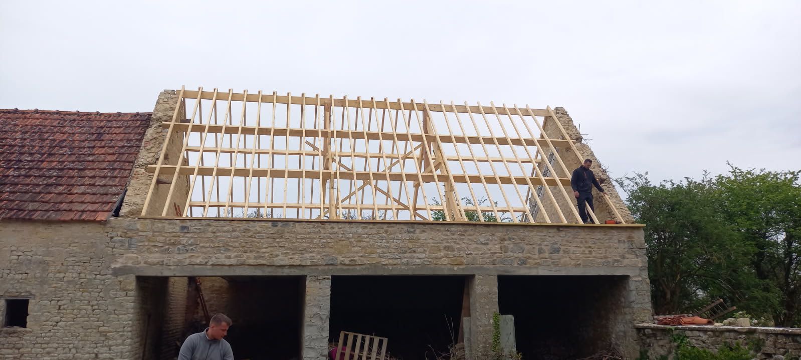 Des ouvriers construisent un toit sur un bâtiment en pierre. Le ciel est nuageux.