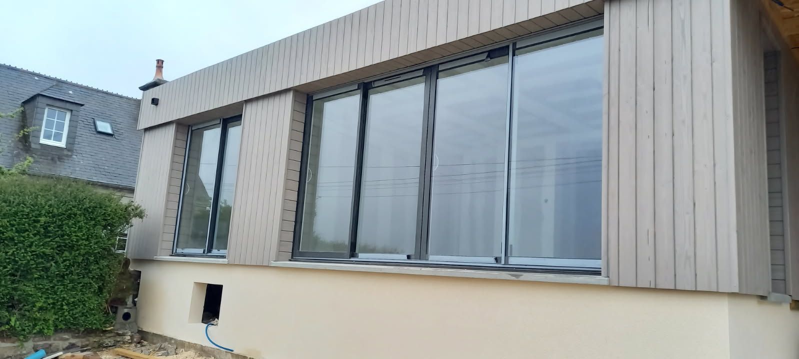 Bâtiment moderne avec de grandes fenêtres, un revêtement clair et une petite haie verte.