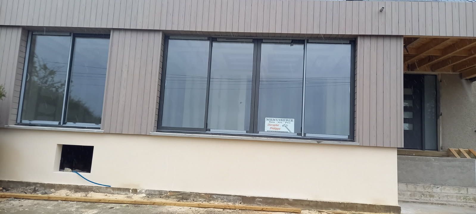 Extérieur d'une maison moderne avec de grandes fenêtres, une porte et un mur beige. Revêtement en bois marron.