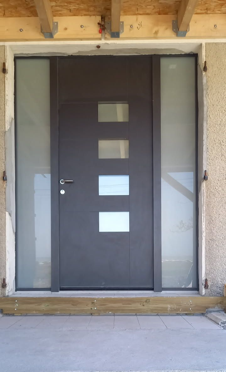 Porte d'entrée grise avec panneaux latéraux givrés et fenêtres rectangulaires. Porche en béton, sous un avant-toit en bois.