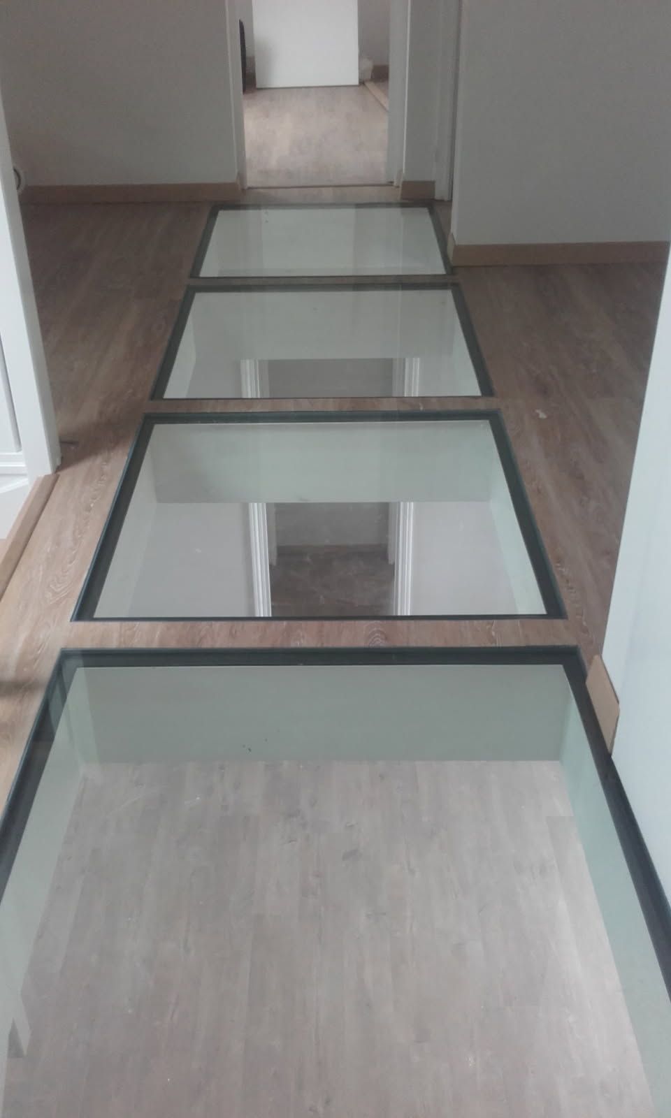 Des panneaux de verre intégrés dans un plancher en bois forment une passerelle dans un couloir, offrant une vue sur l'espace en dessous.