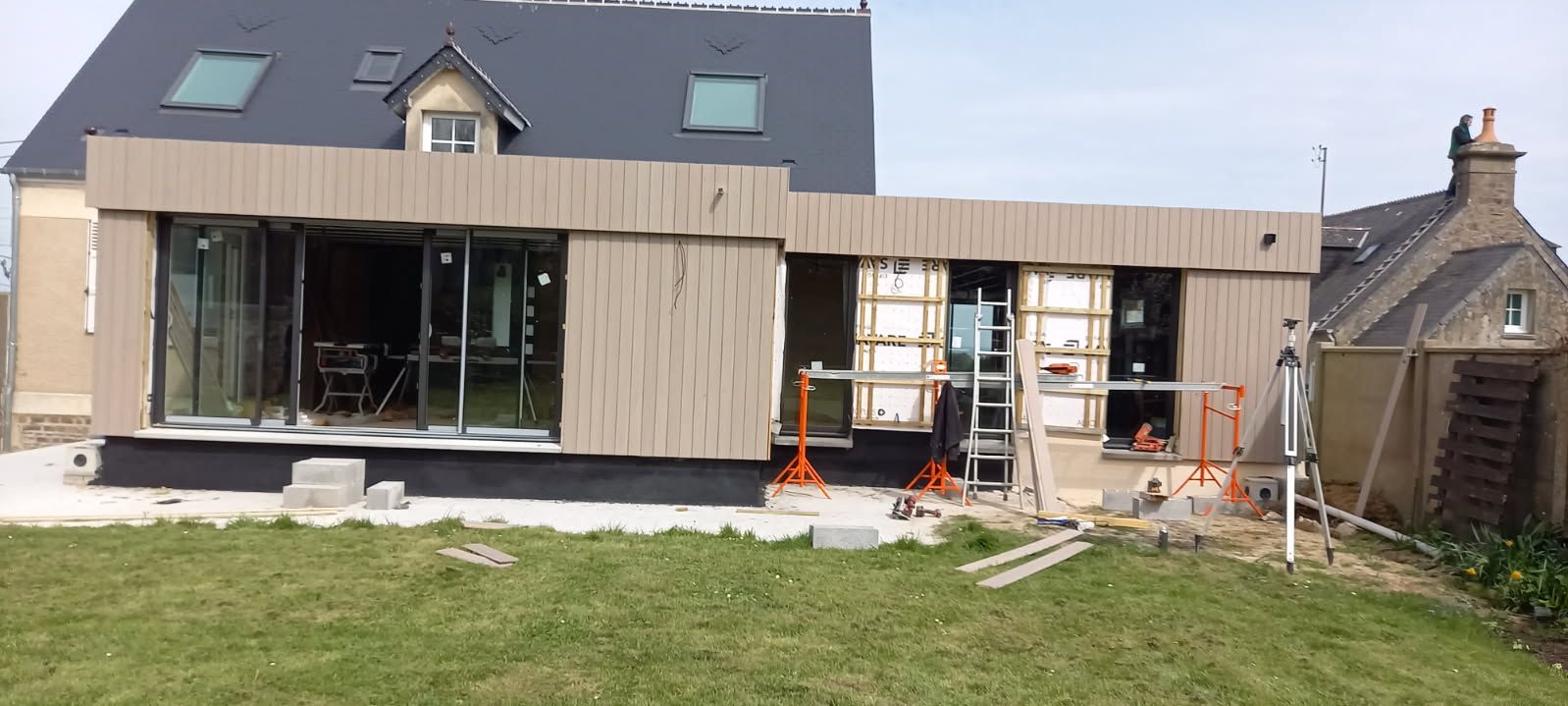 Chantier : extension d'une maison moderne avec façade en bois et grandes baies vitrées. Pelouse verte devant.