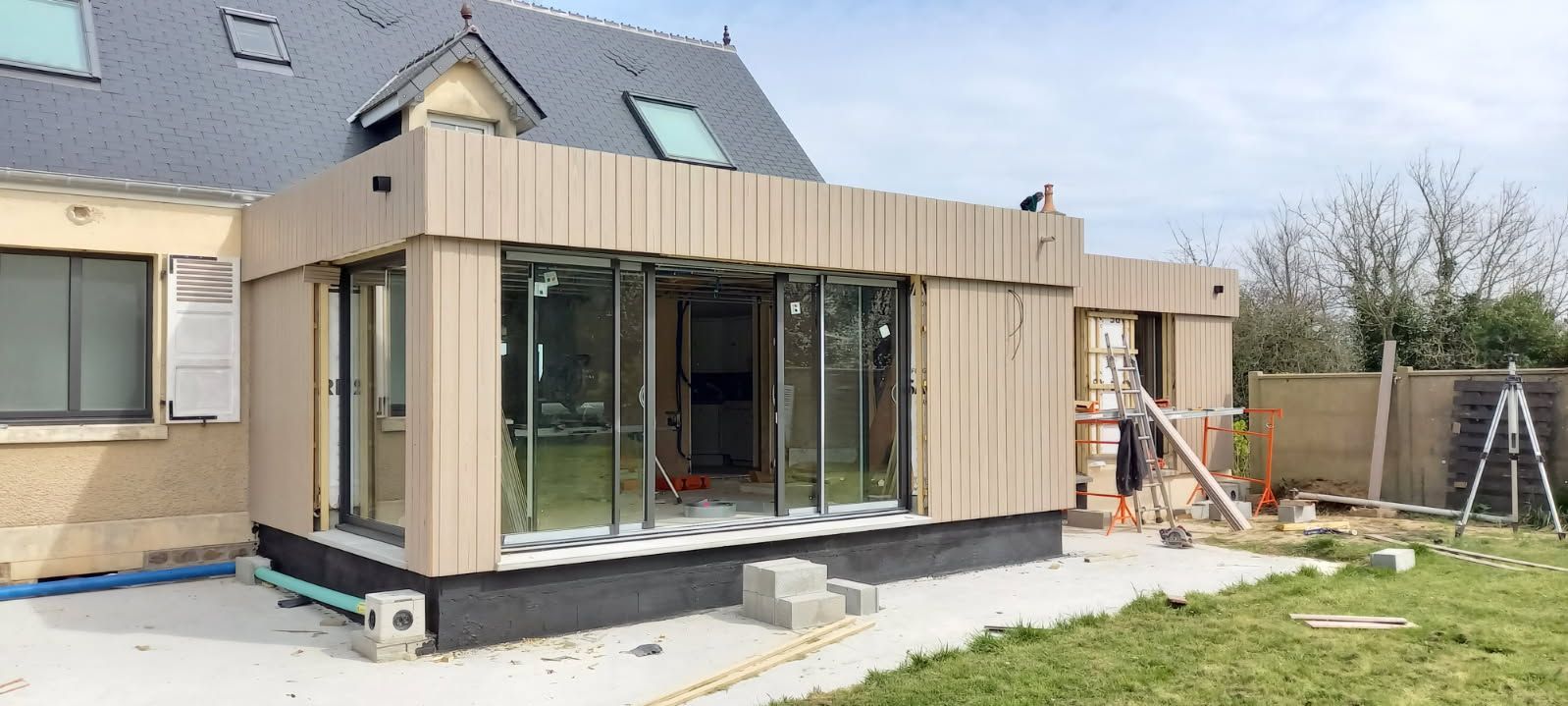 Construction d'une véranda pour une maison; revêtement brun clair, portes coulissantes en verre, pelouse et échafaudage.