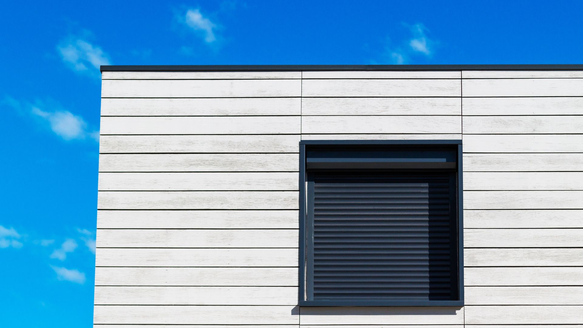 Extérieur d'un bâtiment moderne avec revêtement horizontal blanc, fenêtre bleue avec volet et ciel bleu vif.
