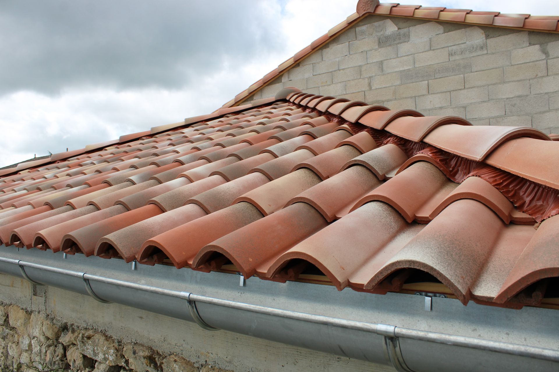 Tuiles en terre cuite rouge avec une gouttière en métal contre un ciel nuageux.