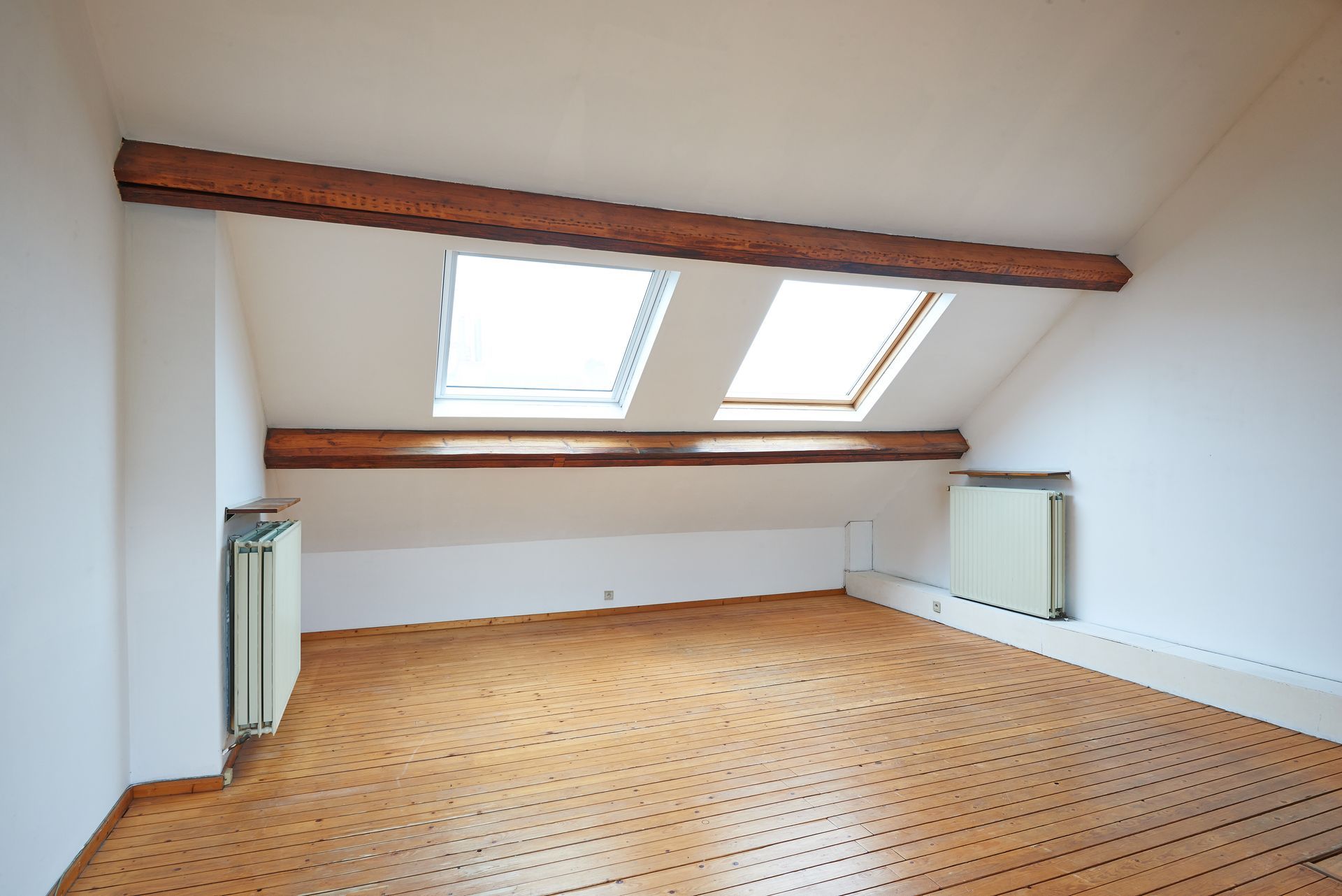 Chambre mansardée avec plancher en bois, plafond mansardé avec puits de lumière, poutres en bois et murs blancs; deux radiateurs.