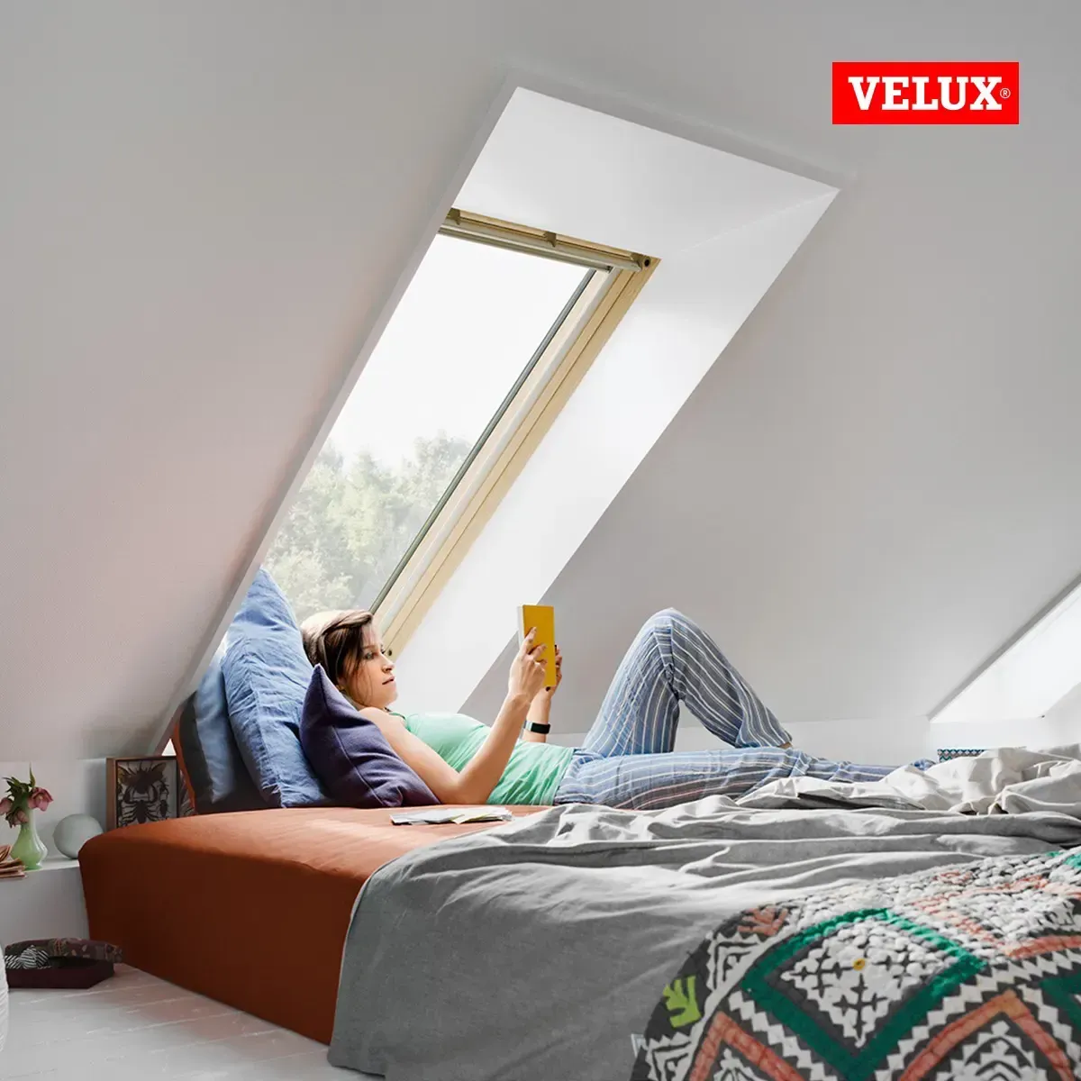 Una mujer acostada en una cama leyendo un libro con un logotipo de Velux en la esquina.
