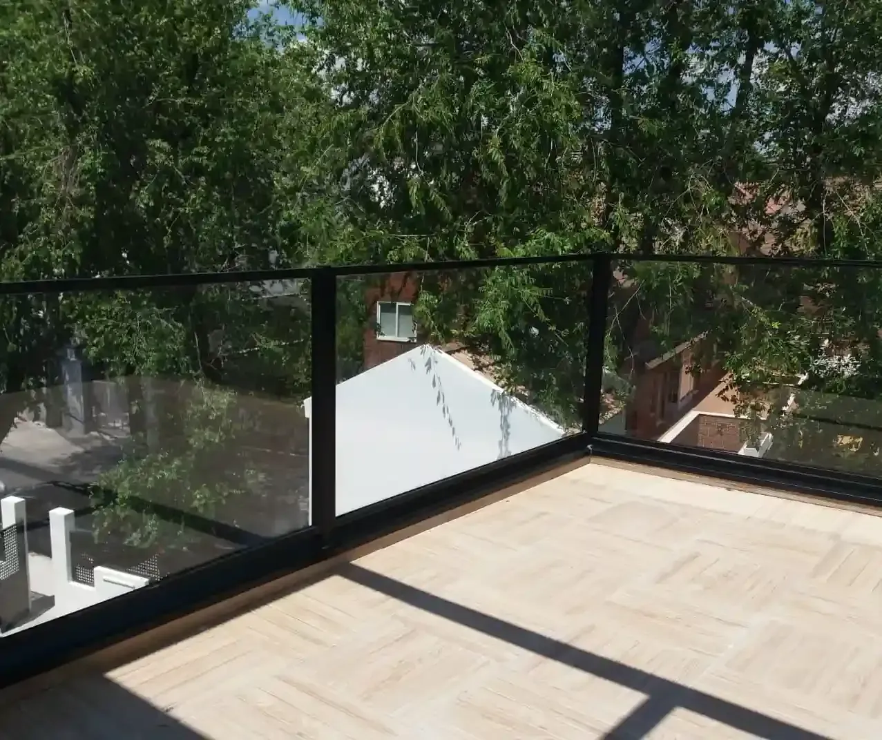 Un balcón con barandilla de cristal y árboles al fondo.