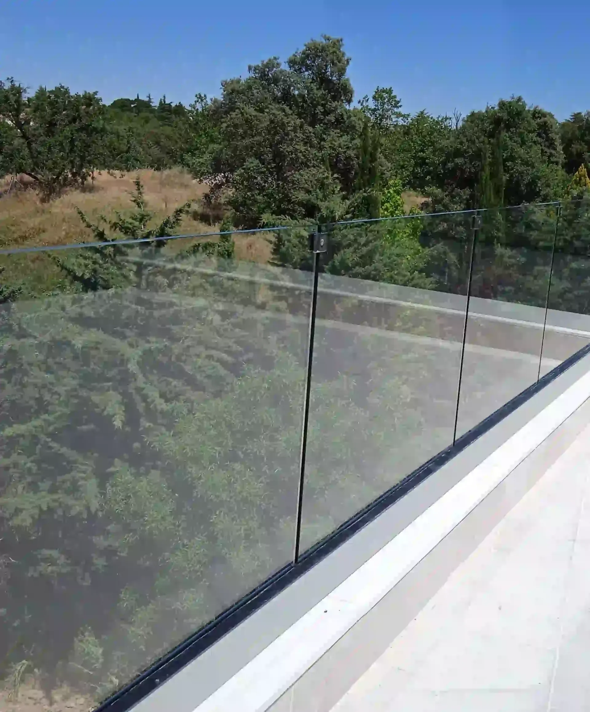 Un balcón con barandilla de cristal y árboles al fondo.