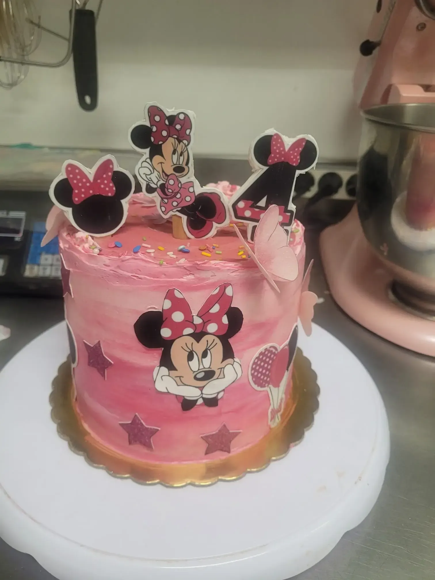 Tarta de cumpleaños rosa de Minnie Mouse con adornos de estrellas y figuras de los personajes, sobre un soporte blanco para tartas