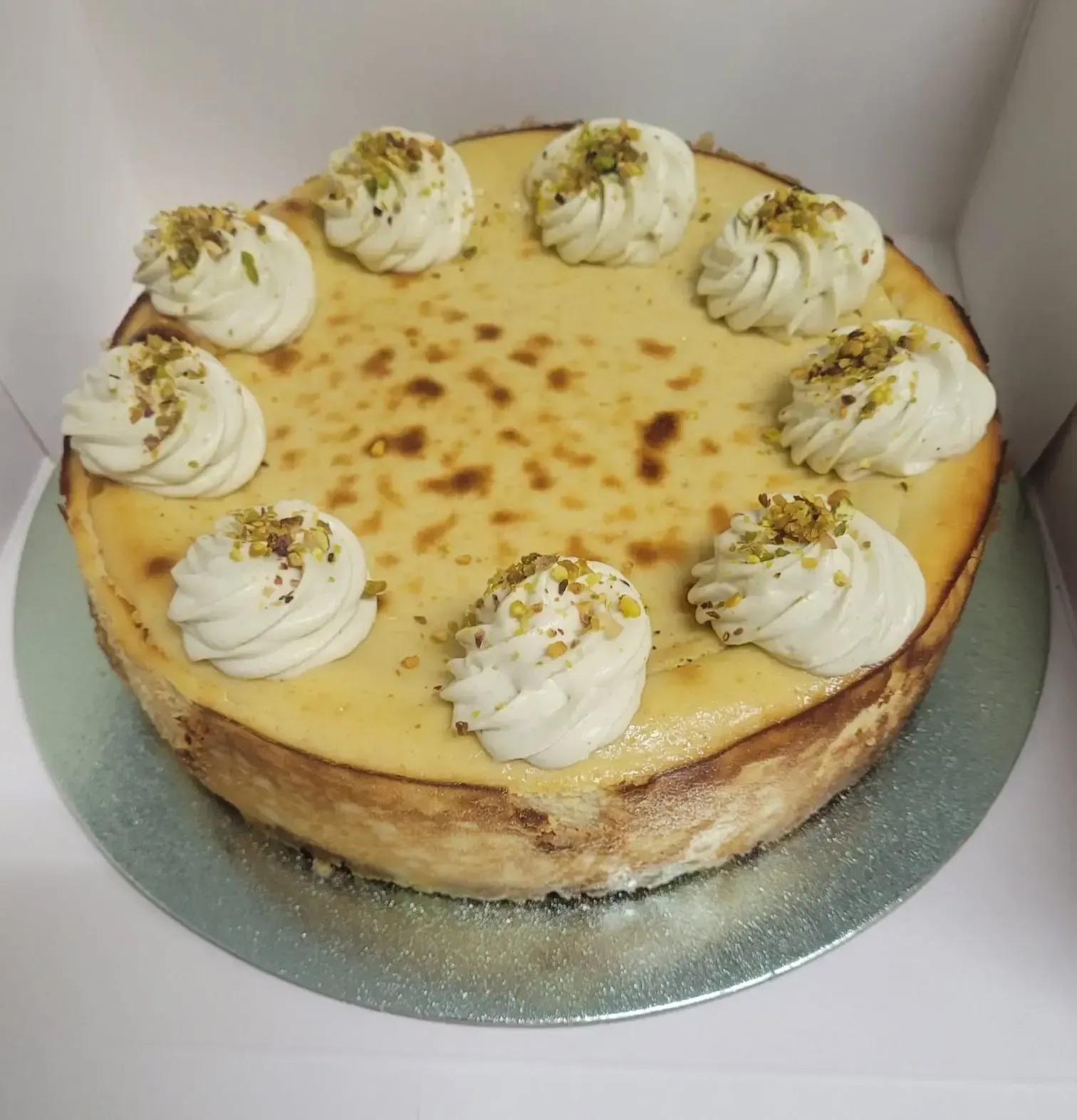 Tarta de queso redonda cubierta con rosetones de nata montada y frutos secos picados, sobre una base plateada para tartas