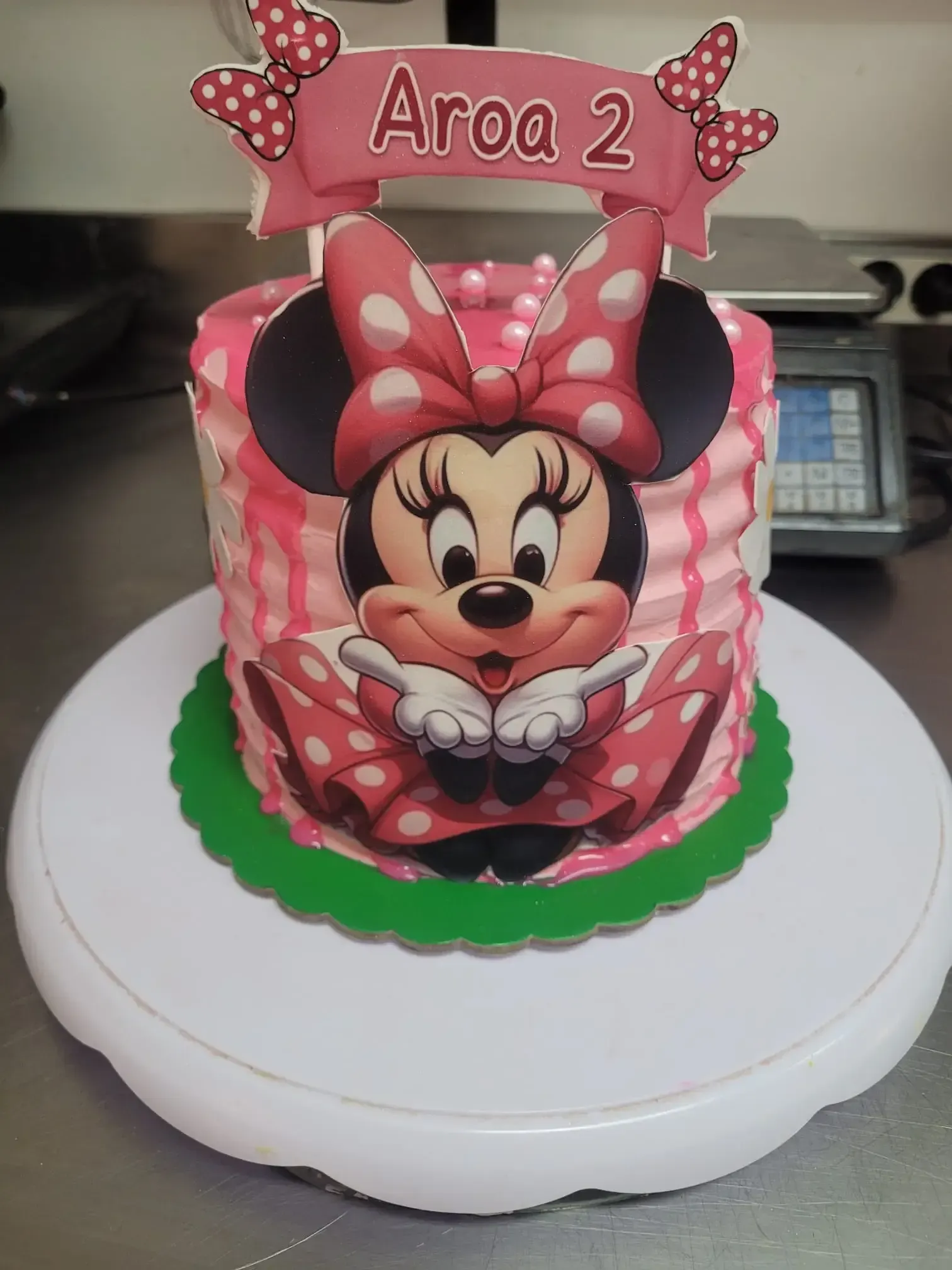 Pastel de cumpleaños de Minnie Mouse con un lazo rosa, con la etiqueta «Aroq 2», sobre un soporte blanco para tartas