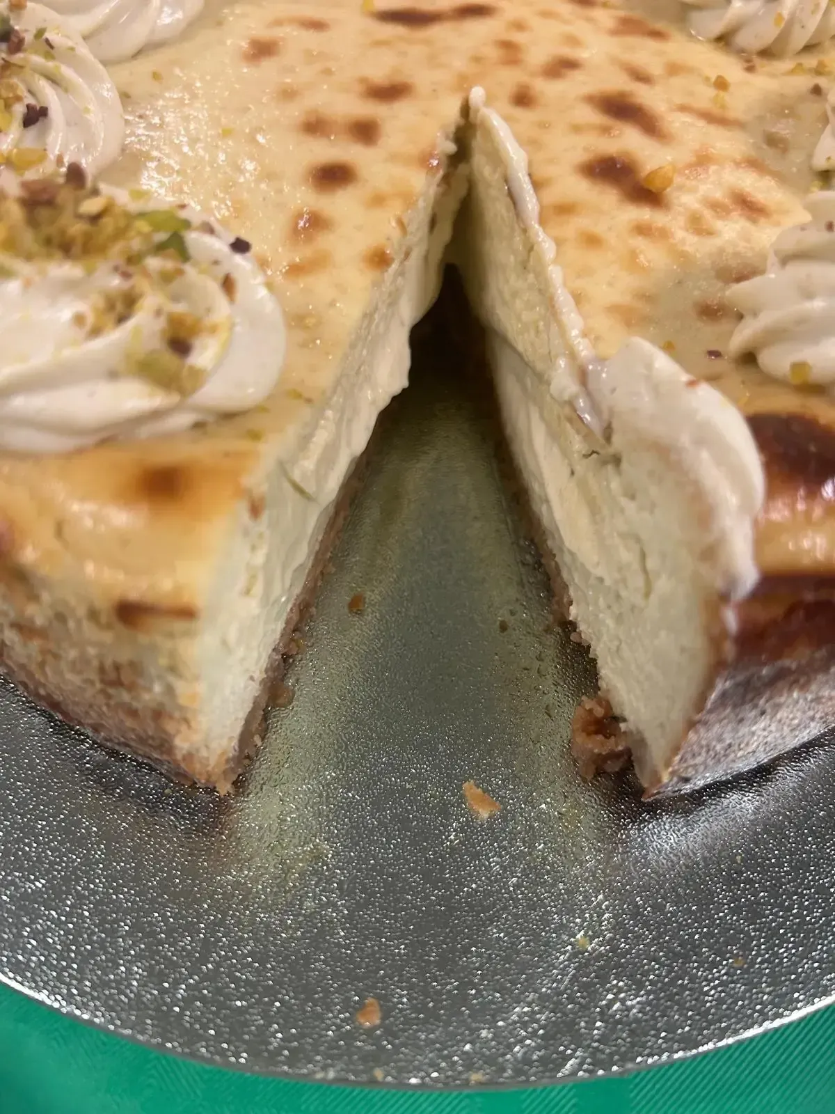 Porción de tarta de queso cremosa con nata montada y pistachos sobre una base plateada para tartas.