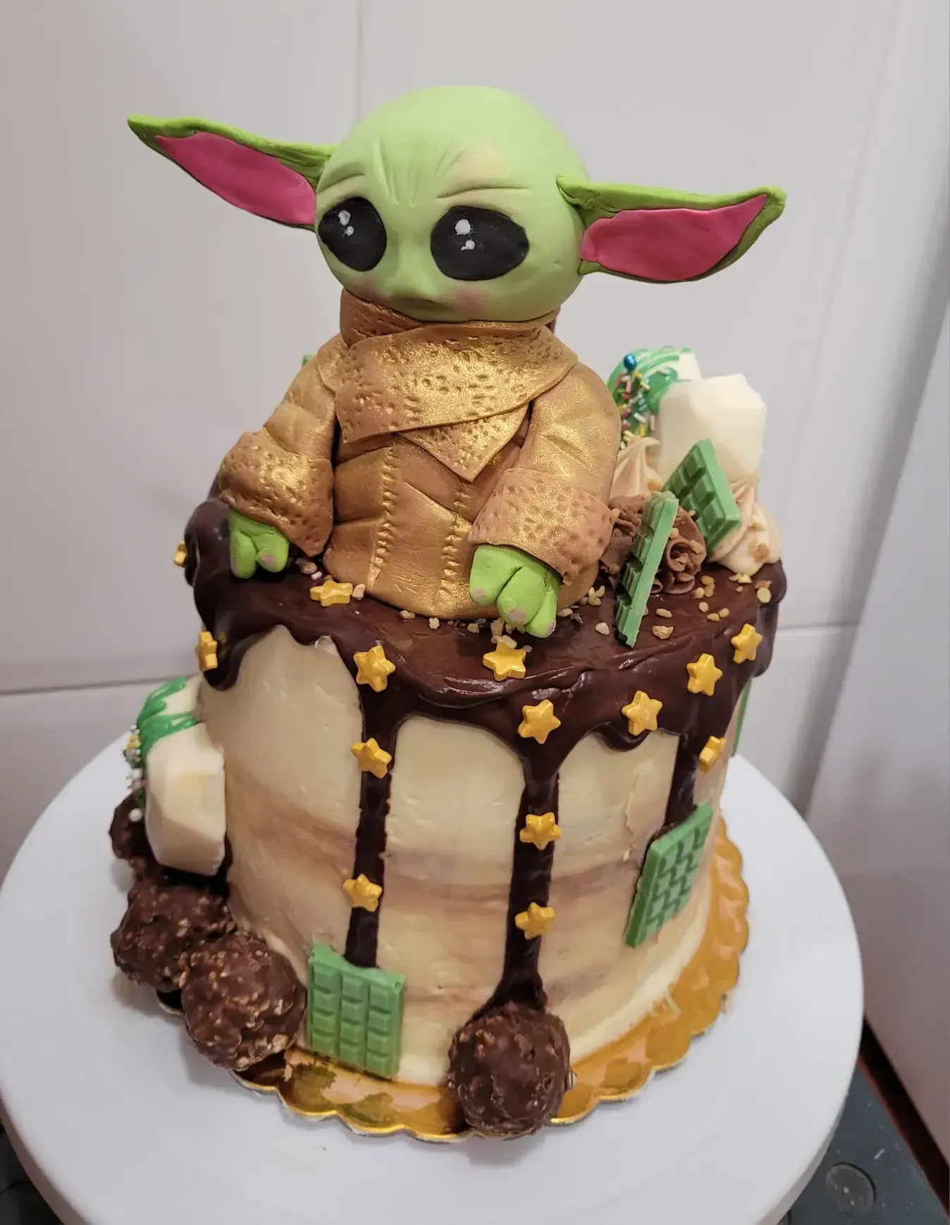 Pastel de Baby Yoda con cobertura de chocolate, detalles verdes y adornos de caramelo sobre una base blanca