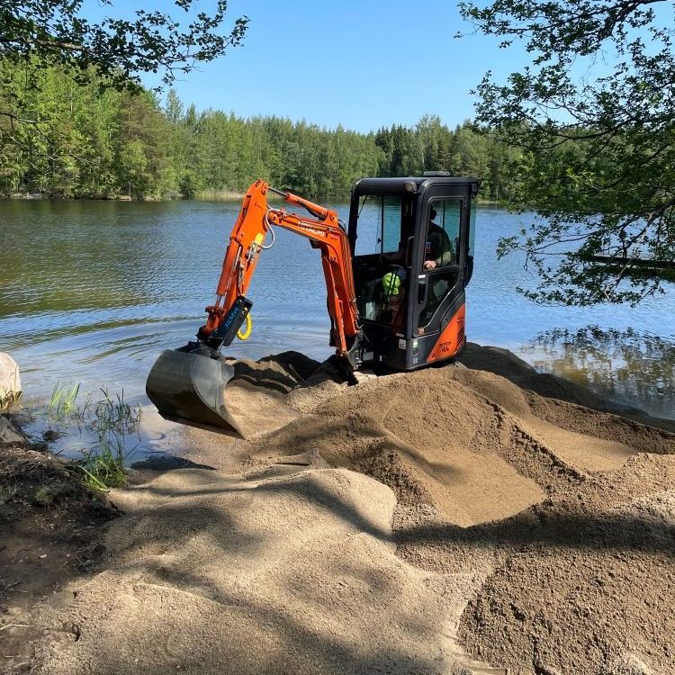 Pieni oranssi kaivinkone kaivaa hiekkaa lähellä järveä
