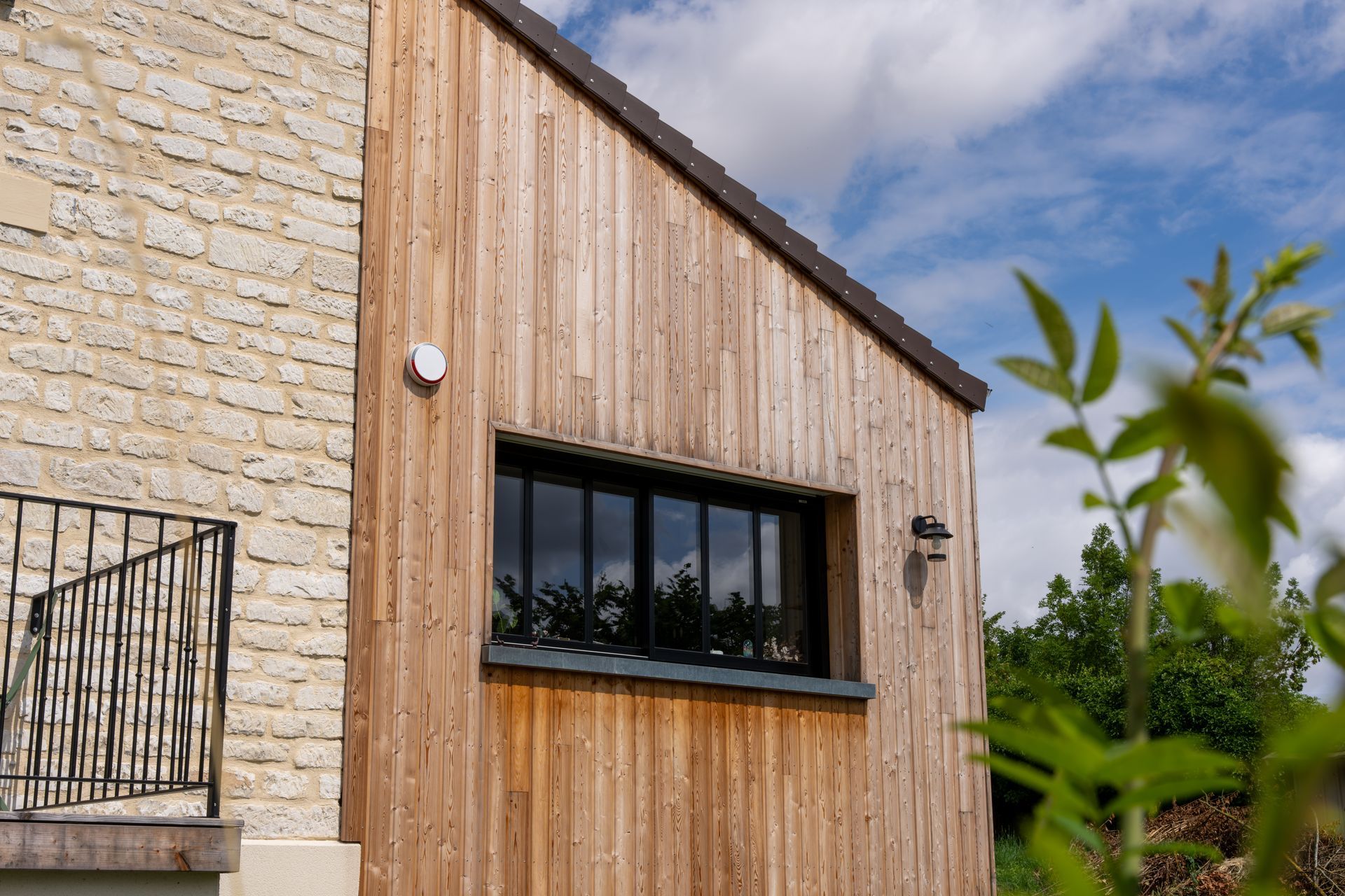 Extérieur d'une maison moderne avec mur en pierre, bardage vertical en bois et fenêtre à cadre noir sous un ciel bleu