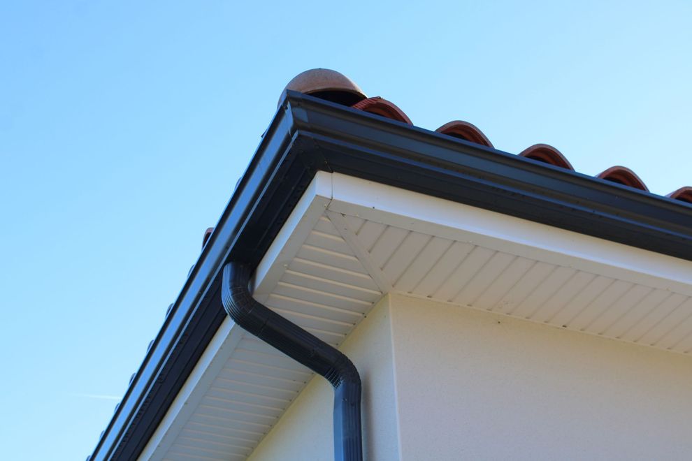 Gros plan sur un coin de toit de maison avec gouttière et descente pluviale noires sur fond de ciel bleu