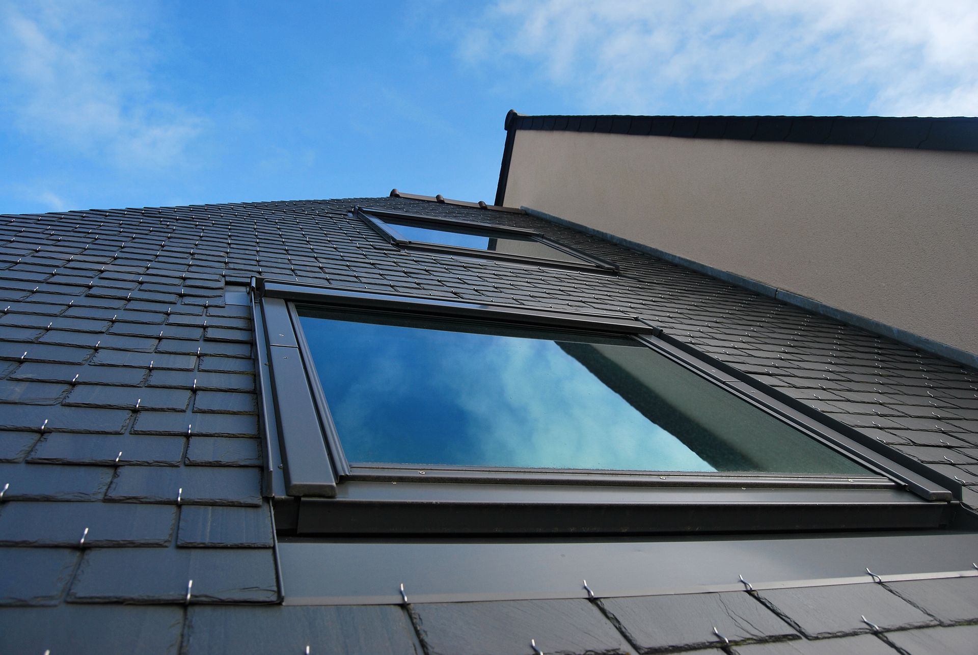 Vue en contre-plongée du toit d'une maison en ardoise sombre, percé d'une lucarne reflétant le ciel bleu.