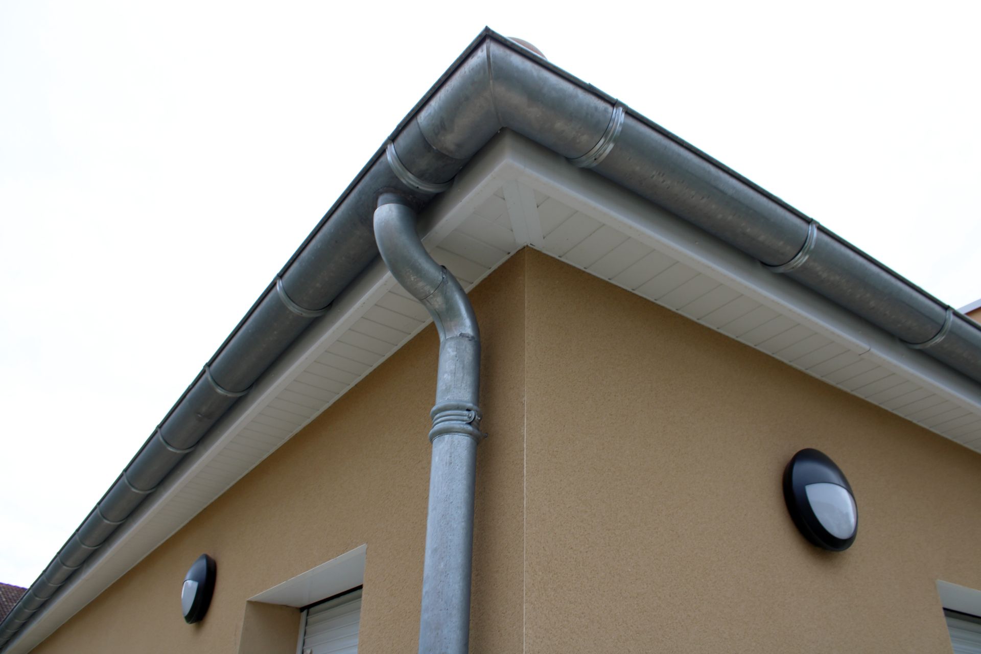 Coin d'une maison beige avec gouttières grises et descente d'eau pluviale sous l'avant-toit.