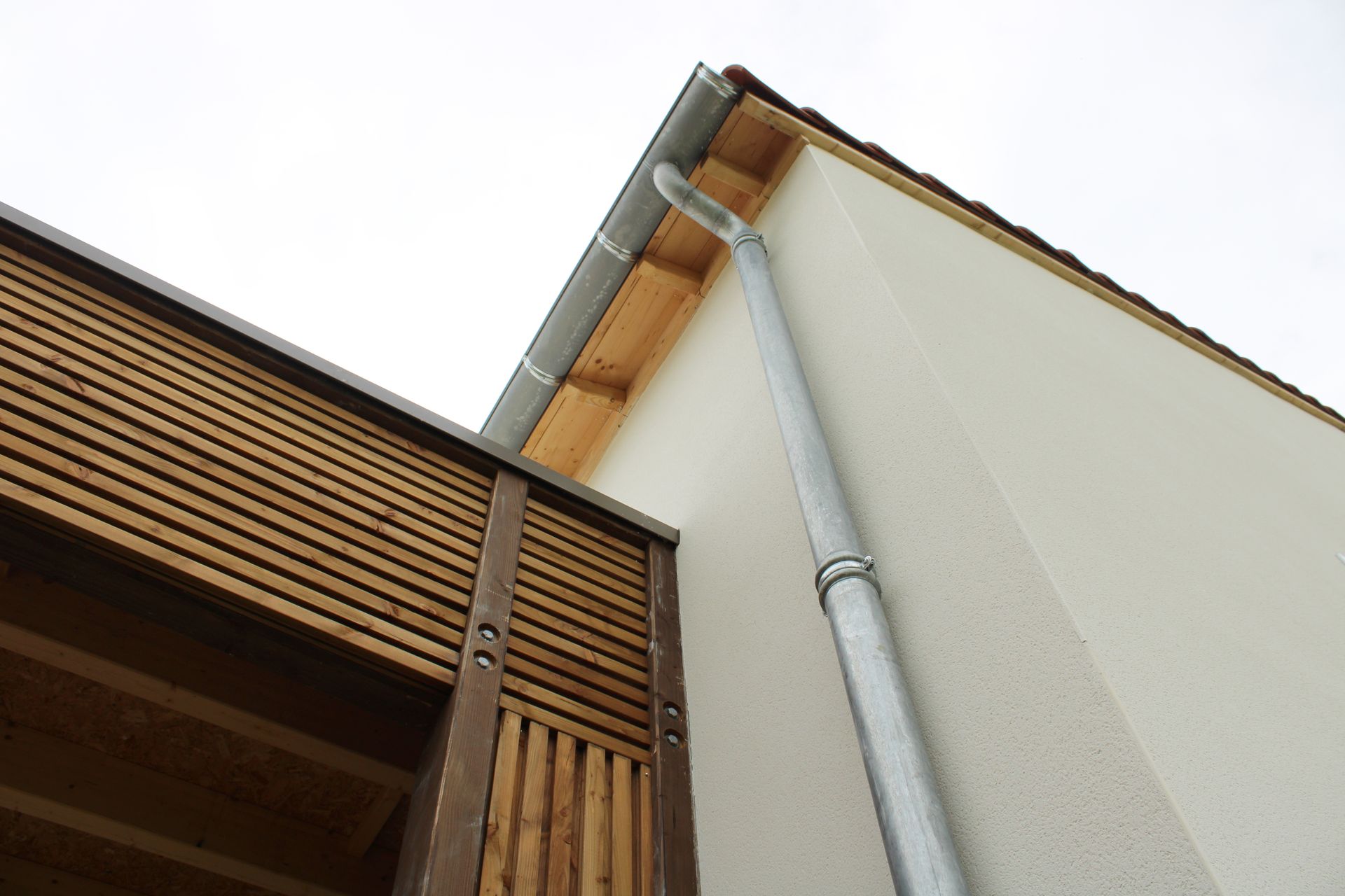 Vue en contre-plongée d'un angle de bâtiment avec un mur en stuc blanc, une descente de gouttière en métal et un bardage en lattes de bois.