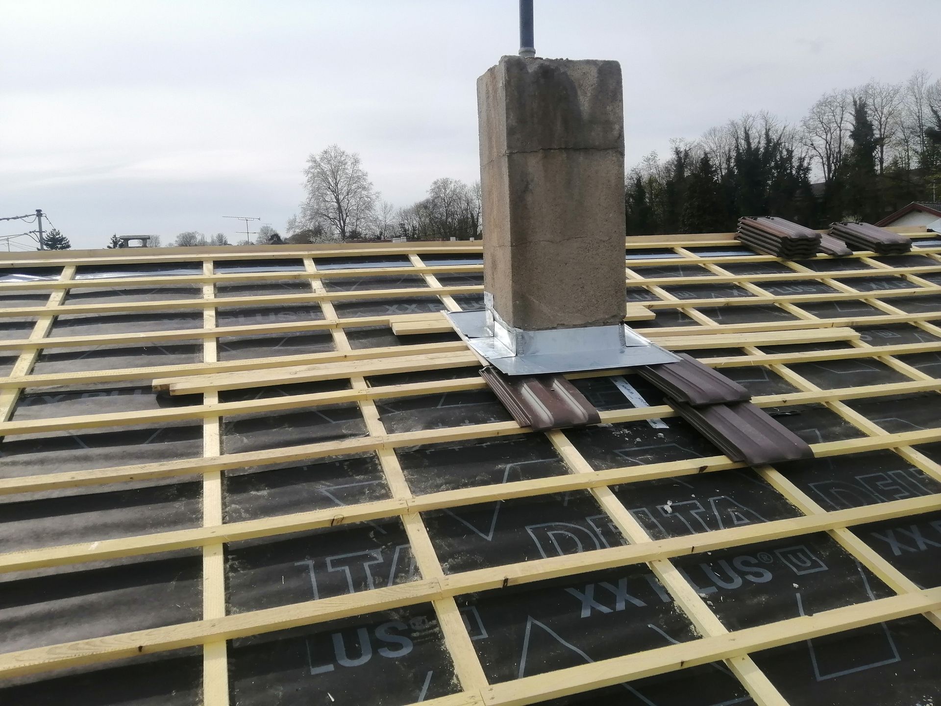 Toiture en construction avec liteaux jaunes, sous-couche noire et cheminée en briques près du faîtage.