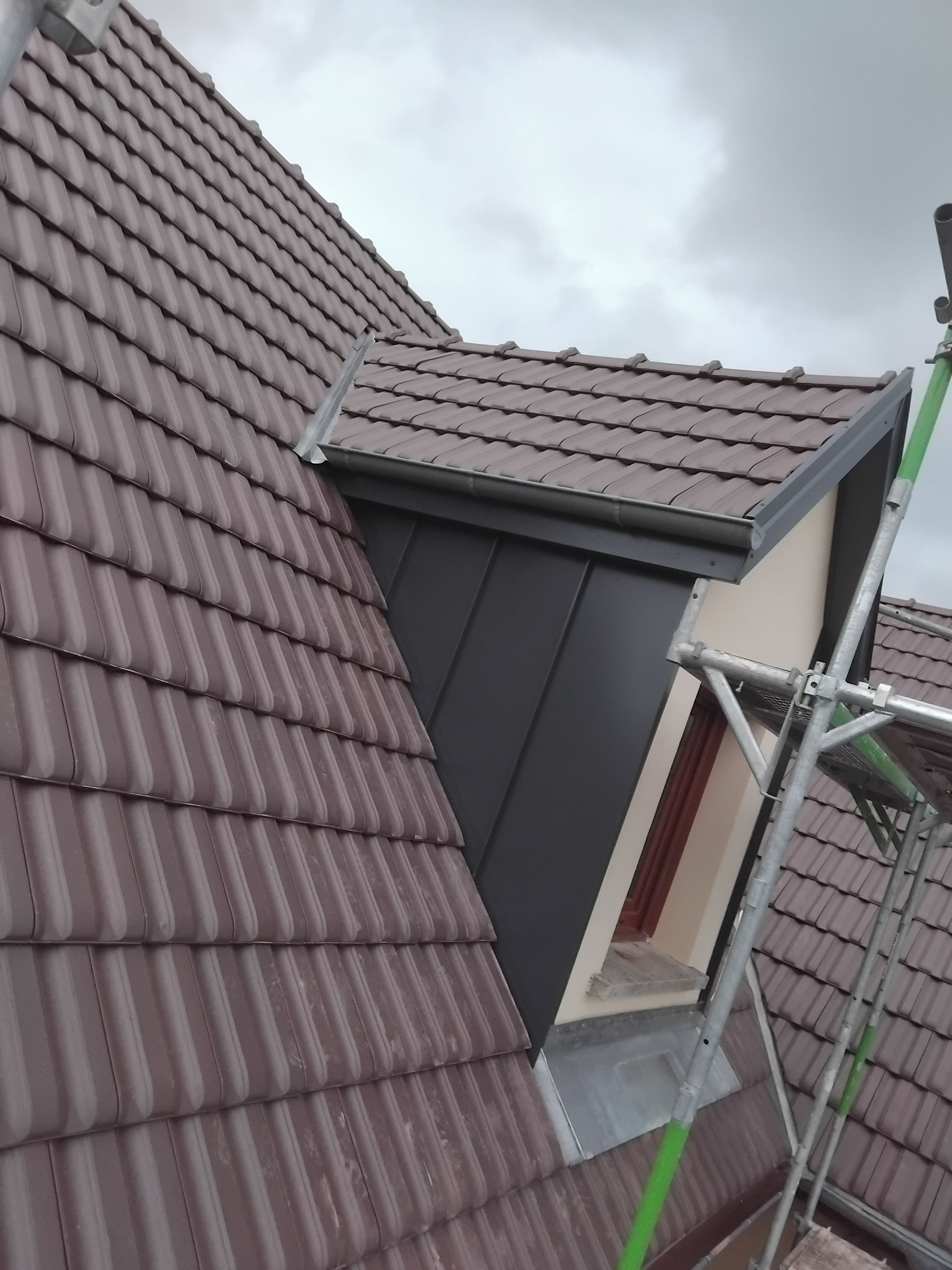Vue inclinée d'un toit de tuiles rouges avec lucarne et échafaudage métallique sous un ciel nuageux