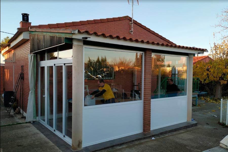 Terraza acristalada adosada a una casa de ladrillo, con paneles de vidrio y molduras blancas. Persona visible en el interior.