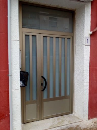 Puerta de metal color canela con paneles de vidrio y ventana lateral, manija negra y buzón, ubicada en la fachada del edificio en blanco y rojo.
