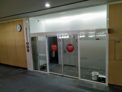 Entrada a baño público con puertas corredizas automáticas de vidrio, paredes blancas y reloj.