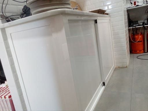 Mueble de cocina blanco con puertas correderas y encimera. Bombona de gas al fondo.