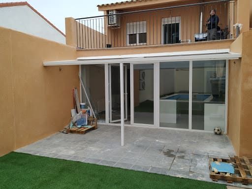 Terraza acristalada con marcos blancos, con piscina visible en el interior. Paredes naranja pálido, césped verde y balcón en el segundo piso.