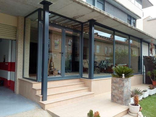 Terraza acristalada adosada a un edificio, con escalones que conducen a la entrada. Marco gris, azulejos color canela, plantas visibles.