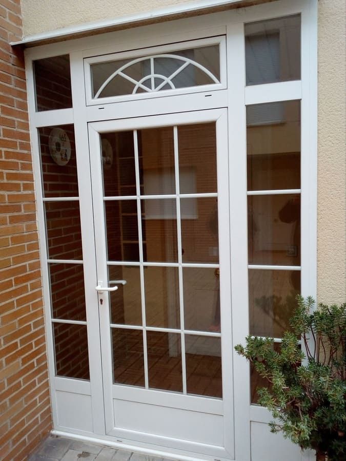 Puerta de vidrio con marco blanco, ventanas laterales y travesaño arqueado, en un exterior de edificio de ladrillo y beige.