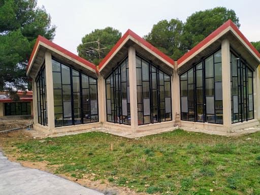 Edificio de cristal de forma triangular con techo rojo, situado en una zona verde con árboles al fondo.