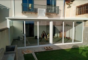 Patio acristalado con marcos blancos, junto a un edificio con balcón. Interior con materiales de construcción.