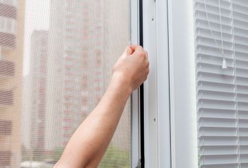 Persona abriendo una ventana blanca con mosquitera; se ven las persianas.