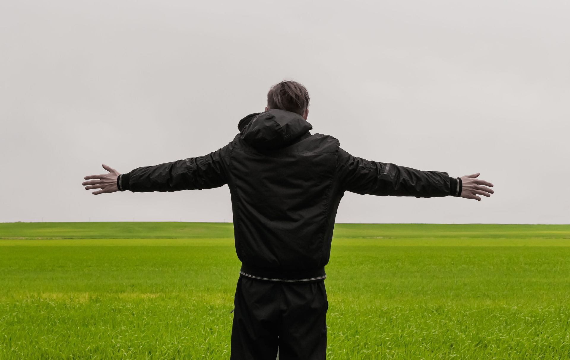 Una persona con una chaqueta negra se encuentra en un campo verde con los brazos extendidos bajo un cielo nublado.