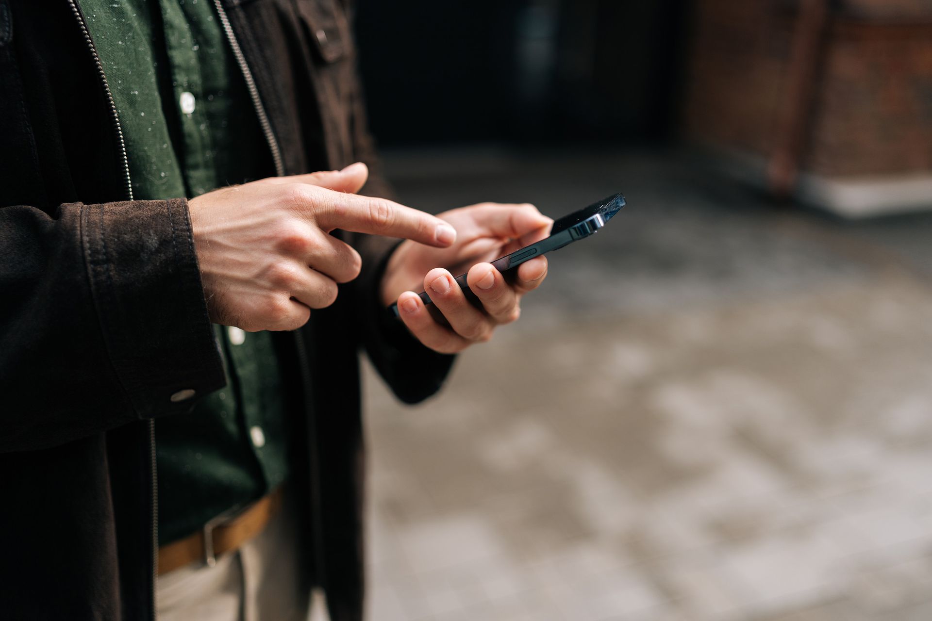 Una persona con una chaqueta marrón usa un teléfono inteligente al aire libre, con el dedo índice tocando la pantalla.