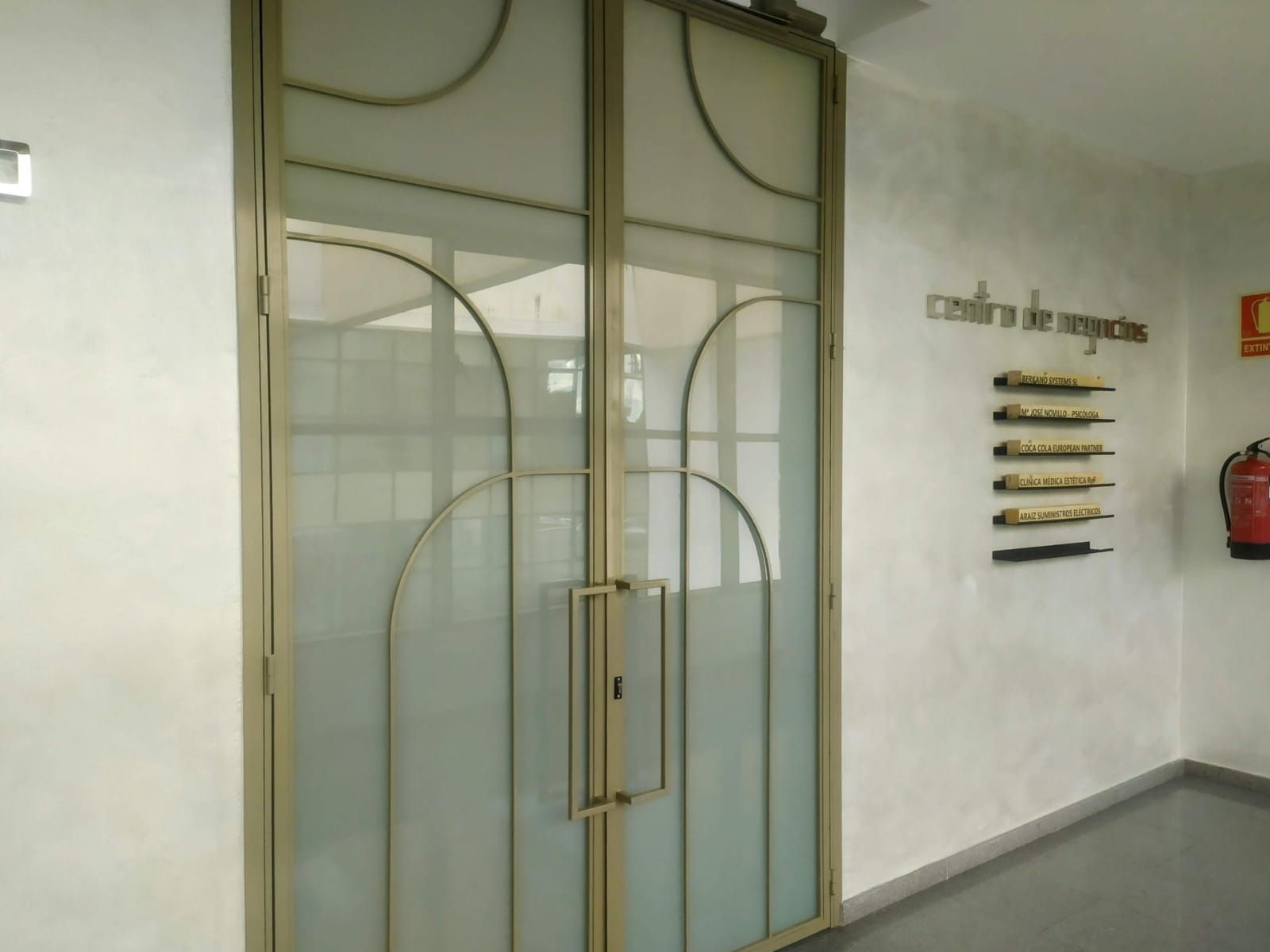 Puertas dobles de vidrio con marco y tiradores metálicos. A la derecha hay un extintor y una decoración de pared.