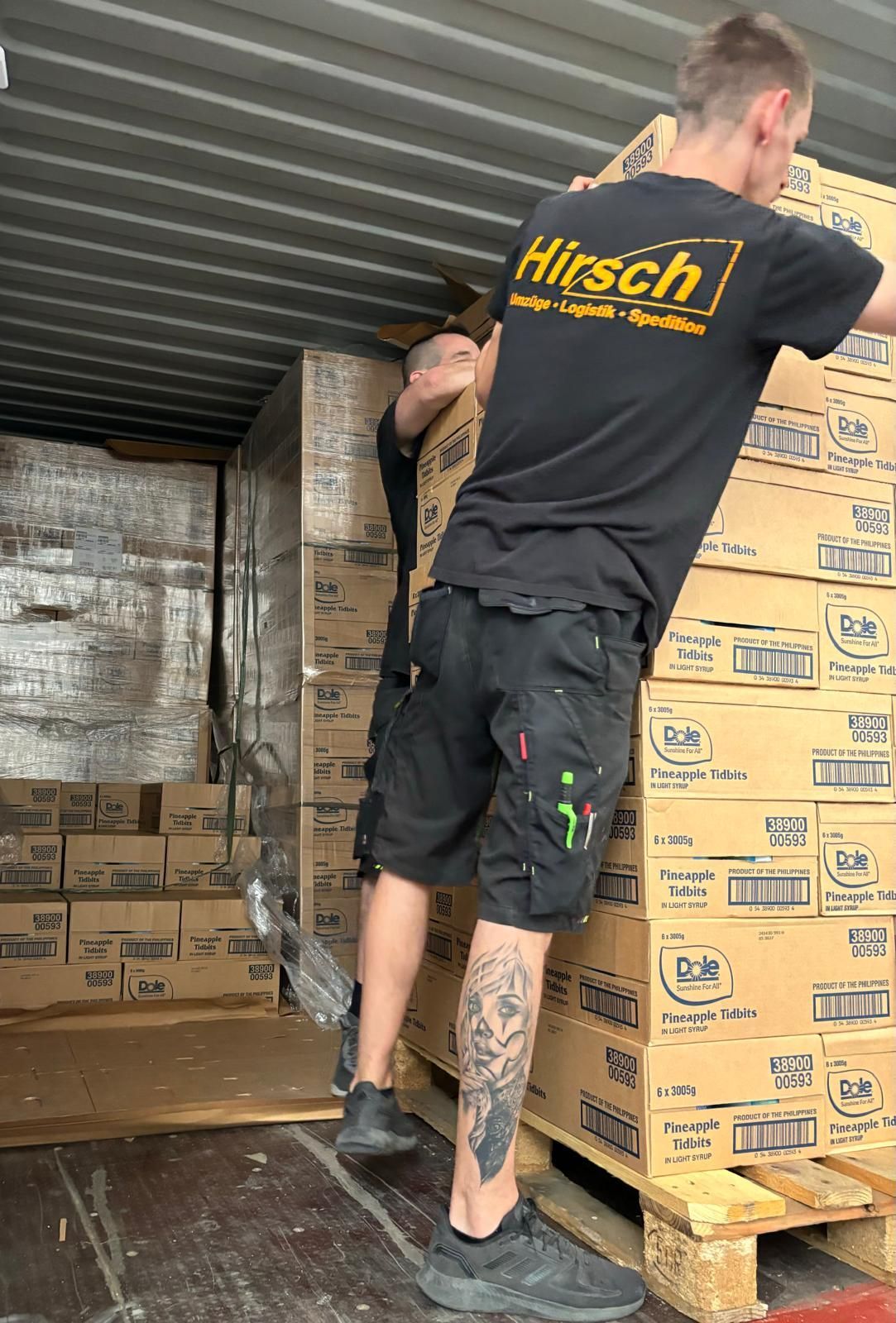 Zwei Männer in schwarzen Hemden und Shorts laden in einem Laderaum Kartons auf einen Lastwagen.