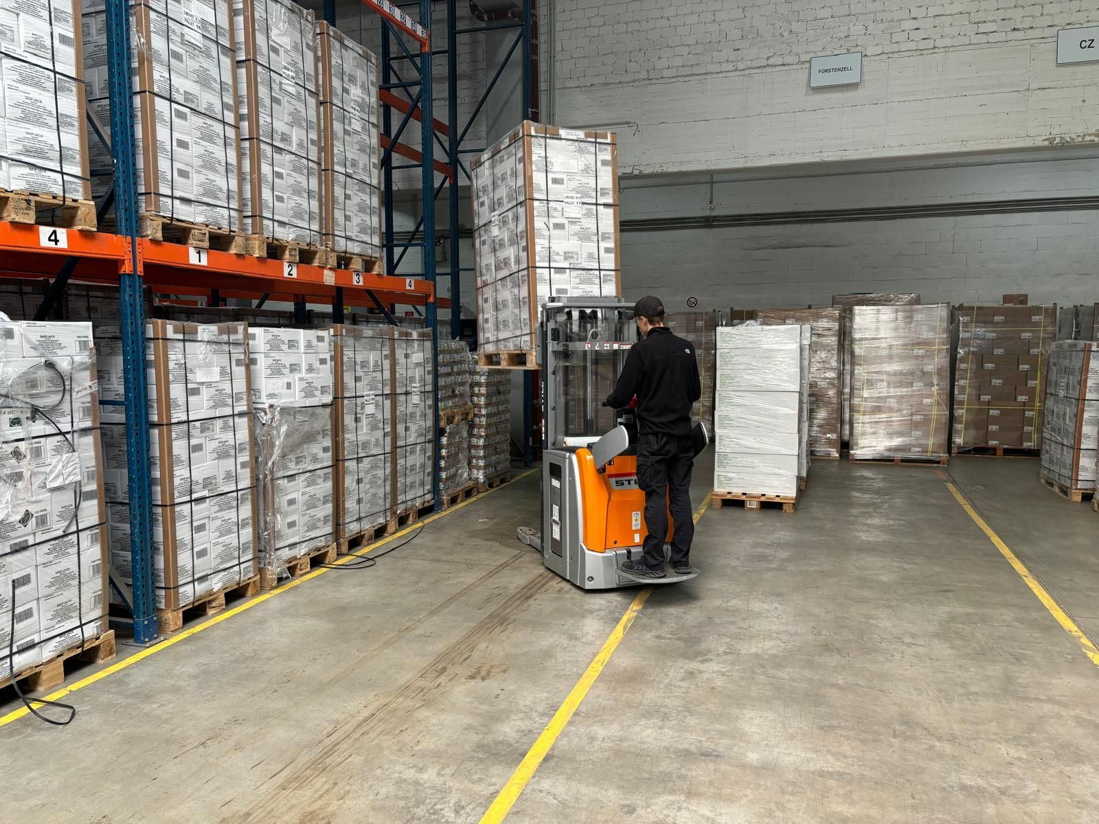 Lagerarbeiter bedient einen Gabelstapler und bewegt Paletten mit verpackten Waren. Reihen gestapelter Kisten.