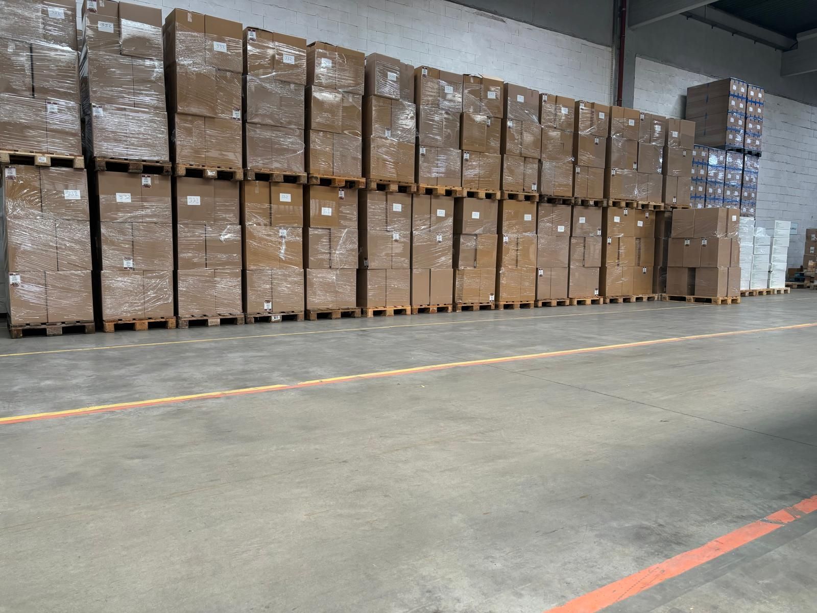 Lager mit gestapelten Kisten auf Paletten; Betonboden mit orangefarbenen Linien.