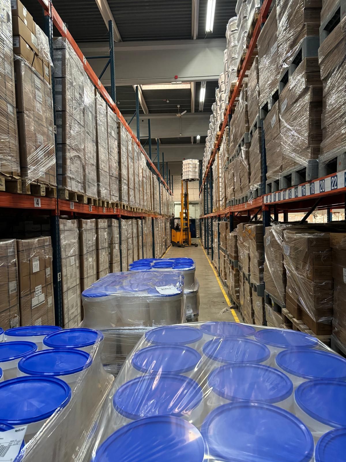 Lagerhalleninneres: Reihen von Paletten mit Waren beladen, blaue Fässer im Vordergrund, Gabelstapler in der Ferne.
