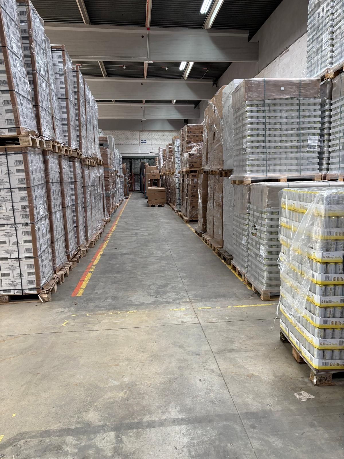 Lagerinnenraum: Stapel verpackter Waren auf Paletten, mit einem zentralen Laufsteg.