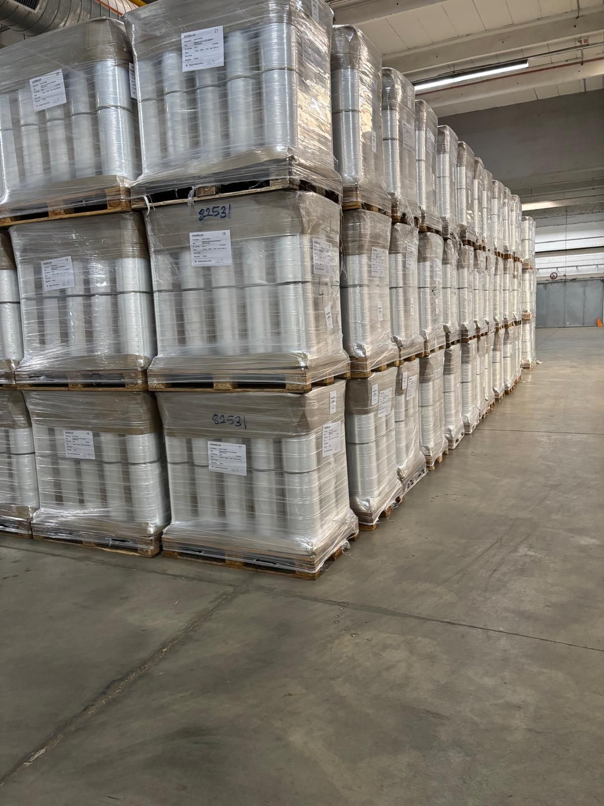 In einem Lager sind Paletten mit zylindrischen, in Plastik eingewickelten Gegenständen gestapelt.