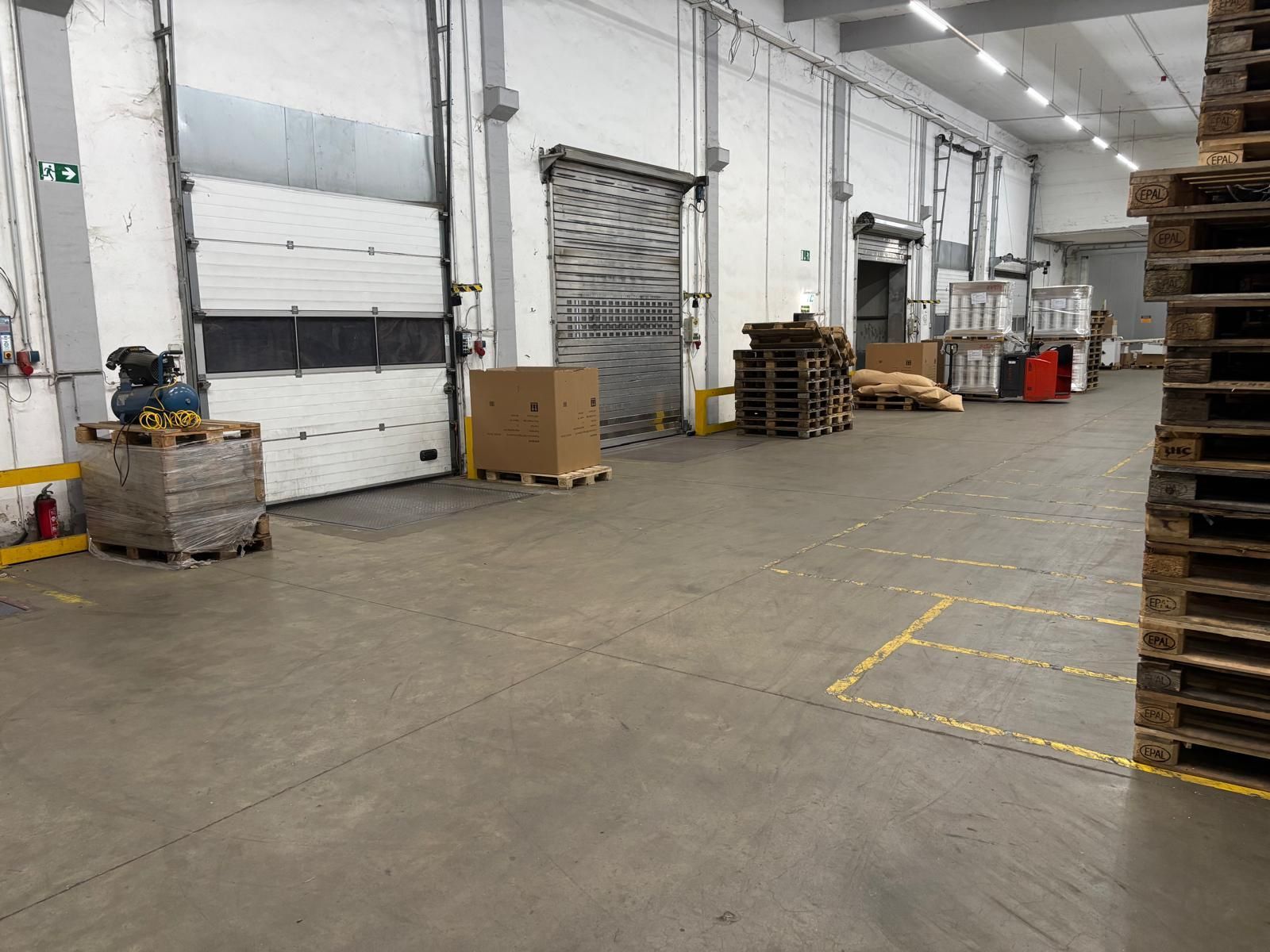 Lagerinnenraum mit Laderampen, Paletten und Kisten. Grauer Betonboden und weiße Wände.