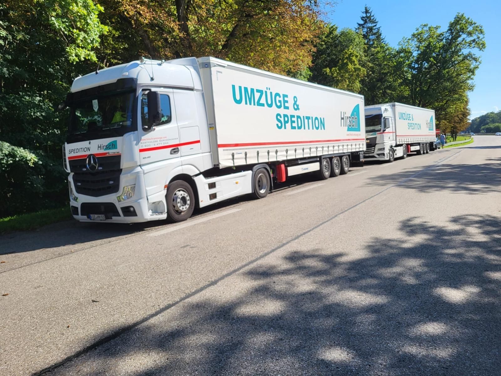 Zwei weiße Sattelschlepper parken auf einer Straße. Auf den Seiten des Anhängers ist in blauer Schrift „Umzüge & Spedition“ aufgedruckt.